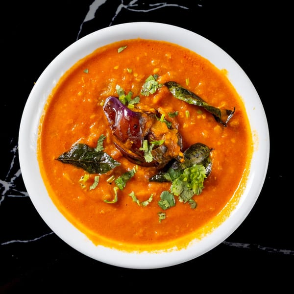 A white bowl of vibrant orange curry with eggplant, curry leaves, and cilantro on a black surface.