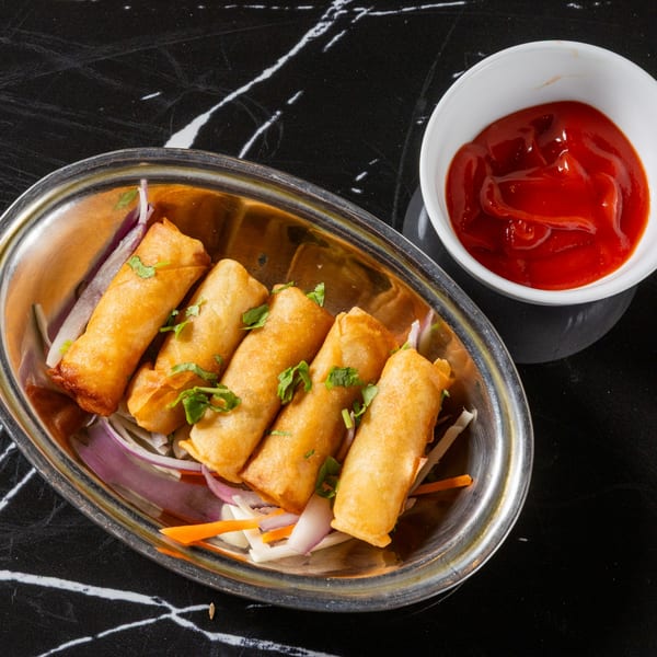 Golden spring rolls with cilantro and shredded vegetables in a silver dish, with ketchup on a black marble table.