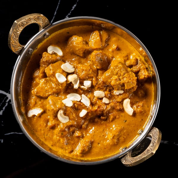 Chicken curry with cashews in a metal bowl with ornate handles on a black marble surface.