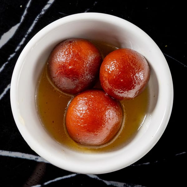Three golden-brown gulab jamun soaked in syrup, served in a white bowl on a dark marble surface.