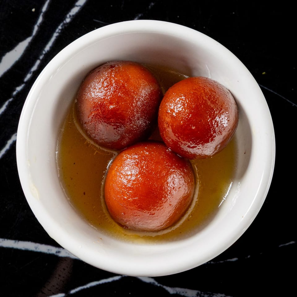 Three golden-brown gulab jamun soaked in syrup, served in a white bowl on a dark marble surface.