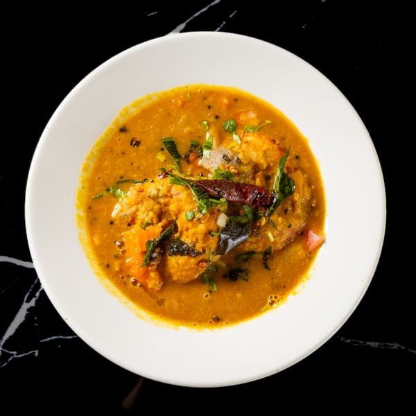 A white bowl of vibrant orange sambar, garnished with herbs and a red chili, on a black marble surface.