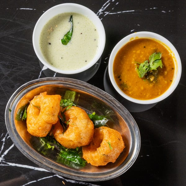 Three golden-fried vada donuts with green herbs in a silver bowl, served with two bowls of dipping sauces.