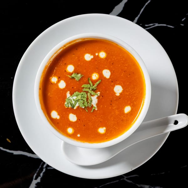 A bowl of vibrant orange soup, garnished with cream swirls and fresh herbs, on a white plate.