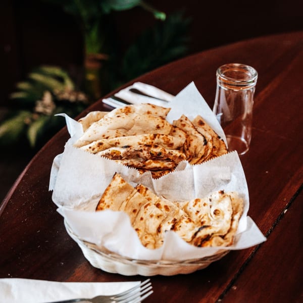 A basket of freshly baked naan bread, lined with white paper, on a dark wooden table.