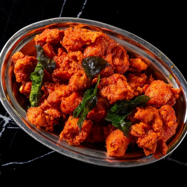 Close-up of bright orange fried chicken pieces garnished with green curry leaves in a silver bowl.